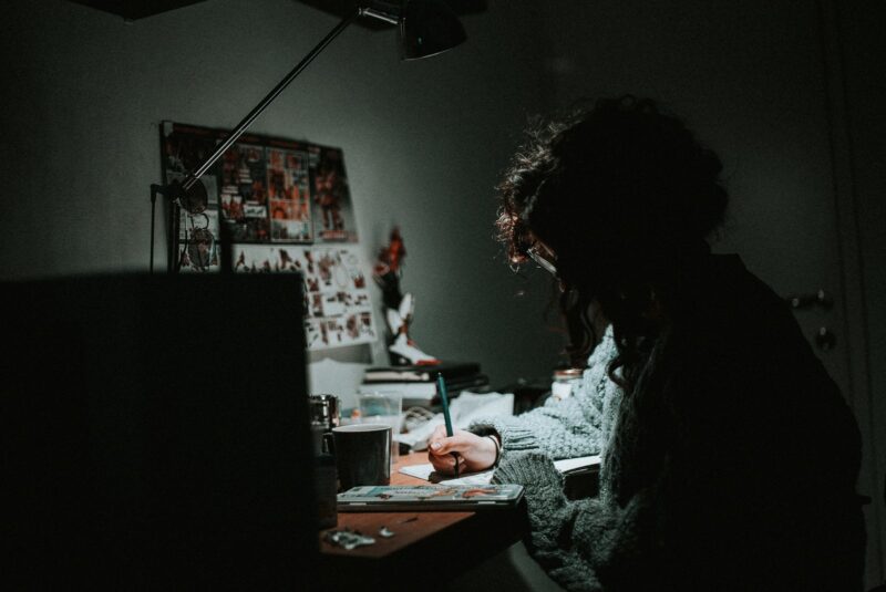 low light photography of woman in gray knit sweatshirt writing on desk